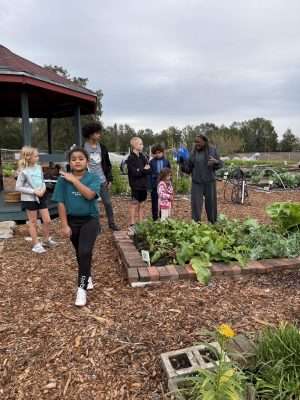 A group of people, including children, stand near a garden bed. An adult is speaking, possibly giving instructions or a tour, with assorted vegetables visible. A gazebo is in the background.